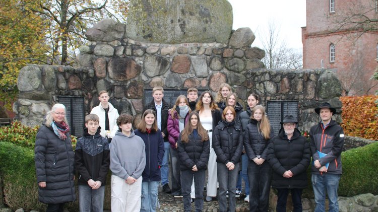 Zu sehen sind Schülerinnen und Schüler die in mehreren Reihen vor einem Gedenkstein stehen und in die Kamera blicken.