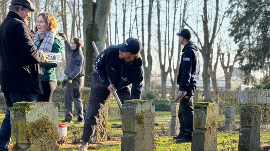 Junge Erwachsene auf einer Kriegsgräberstätte. Einige halten Werkzeuge und Bürsten in der Hand um Reinigungs- und Pflegearbeiten durchzuführen.