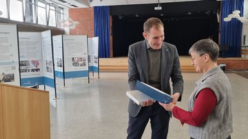 Ein lächelnder Mann übergibt einer Frau eine Urkunde. Links daneben Ausstellungswände