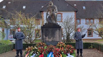 Zwei Männer in Uniform stehen links und rechts neben einem Denkmal, sie halten je eine Fackel in der Hand. Das Denkmal zeigt einen knienden Mann mit Helm, der Kopf ist gebeugt. Vor dem Mahnmal ligen mehrere Kränze.
