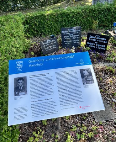 Drei Grabplatten, davor die Informationstafel. Darauf links und rechts je ein Portraitfoto in der Mitte in zwei Spalten der Text.