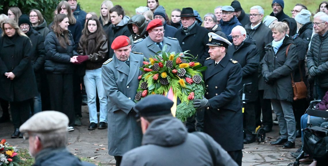 In der Bildmitte tragen drei Soldaten einen Kranz. Um sie steht eine größere Gruppe Menschen.