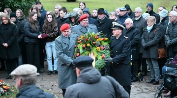 In der Bildmitte tragen drei Soldaten einen Kranz. Um sie steht eine größere Gruppe Menschen.