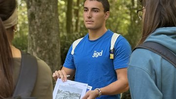 Ein junger Mann hält seine Arbeitsmappe. Er zeigt das Foto einer Person, dessen Biografie er erarbeitet hat. Im Vordergrund sieht man zwei Teilnehmerinnen mit dem Rücken zur Kamera, die dem Jugendlichen und seiner Präsentation zuhören.