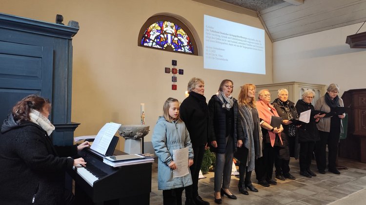 Auf dem Bild ist links eine Frau am Klavier zu sehen. Rechts neben dem Klavier stehen die Sängerinnen im Alter von 8-80 Jahren. 