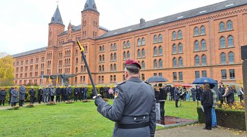 Im Vordergrund (von hinten aufgenommen) ein Mann in Uniform. In einem großen Halkreis darum eine Gruppe Menschen. Im Hintergrund ein großes, altes Gebäude 
