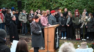 In der Mitte ein Rednerpult hinter dem zwei Schülerinnen stehen. Rechts und links von ihnen Soldaten mit Fakeln. Dahinter ein Mann. Im Hintergrund eine Gruppe Schülerinnen und Schüler.