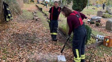 Zwei Personen in Feuerwehruniformen auf einer Kriegsgräberstätte. Die junge Frau reinigt einen Grabstein.