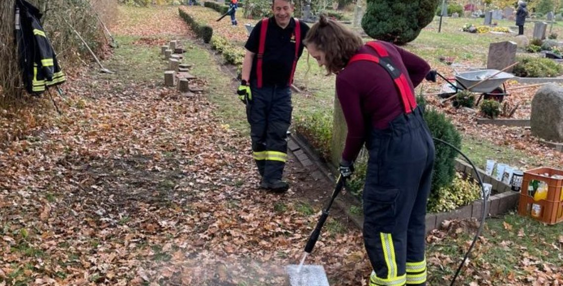 Zwei Personen in Feuerwehruniformen auf einer Kriegsgräberstätte. Die junge Frau reinigt einen Grabstein.